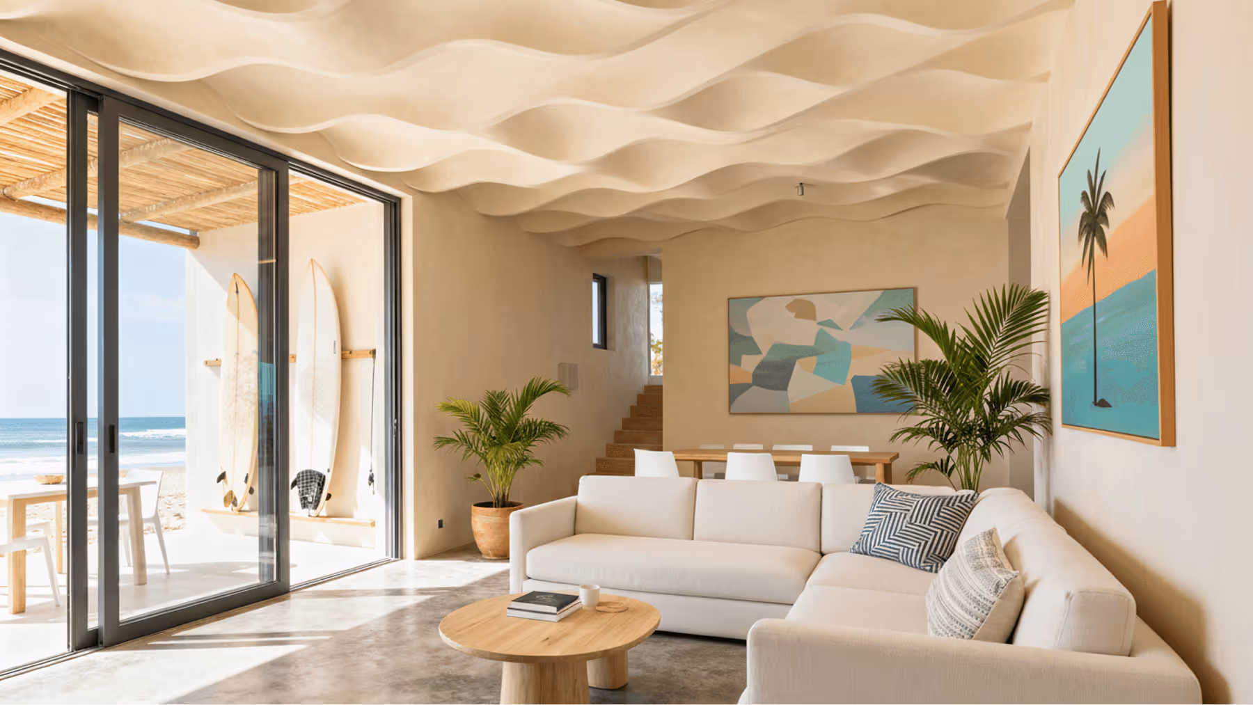 Sala de estar moderna com sofá branco em formato de L, mesa de centro redonda, plantas, duas pranchas de surf na varanda e vista para a praia pelo vidro deslizante.
