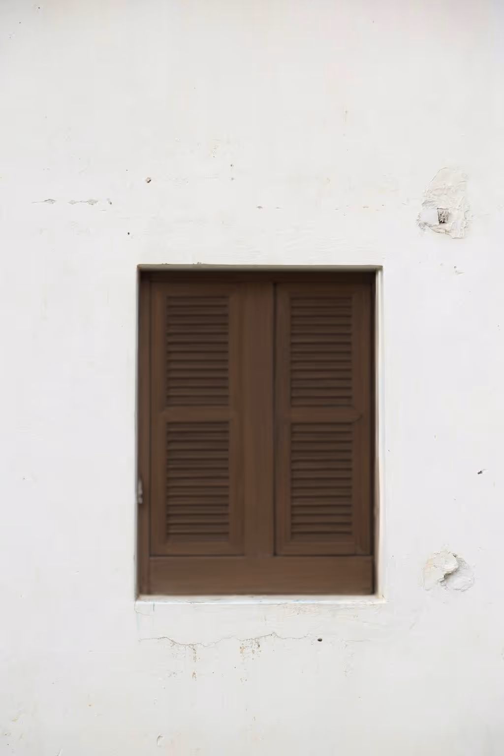 Janela de madeira marrom fechada em uma parede branca com pequenas imperfeições.