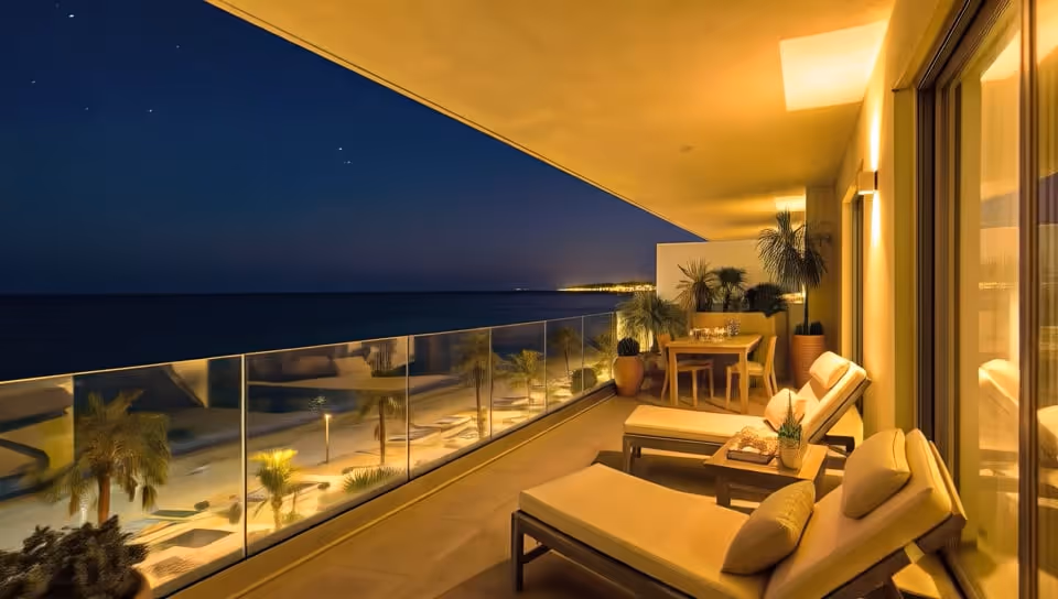 Varanda iluminada à noite com duas espreguiçadeiras, mesa de jantar e vista para a praia e o mar calmo.